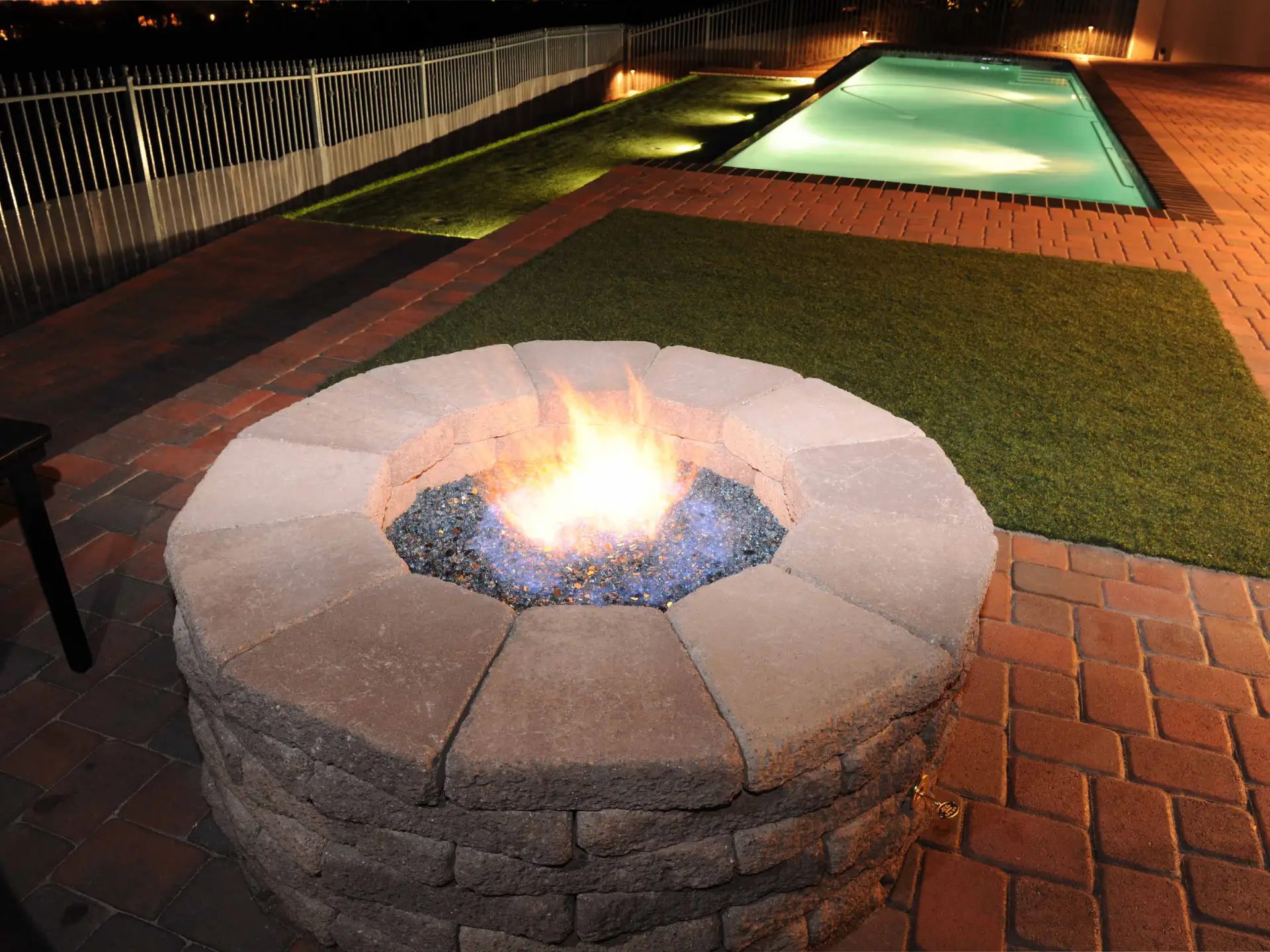 New stone wall fire pit with pool and artificial turf in the background.
