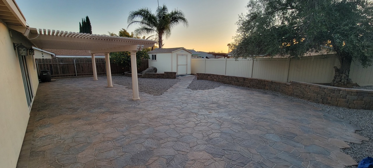 Backyard flagstone patio featuring a curved retaining wall and shade pergola at sunset.